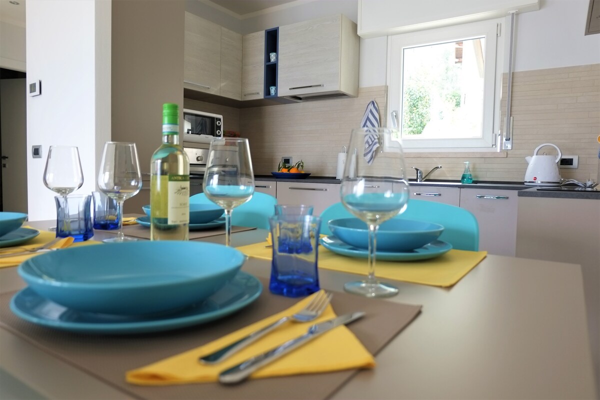 A modern kitchen area showcases organized table settings with blue plates, wine glasses, and yellow napkins. A bottle of white wine is featured, accompanied by a backdrop of cabinets and cooking appliances. Natural light is admitted through a nearby window, illuminating the inviting space.