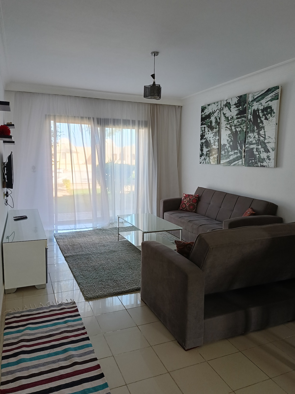 A bright living area features two grey sofas and a glass coffee table, complemented by a striped area rug. Large sliding doors allow natural light to fill the room, revealing views of outdoor greenery. Wall art adds a modern touch to the space.