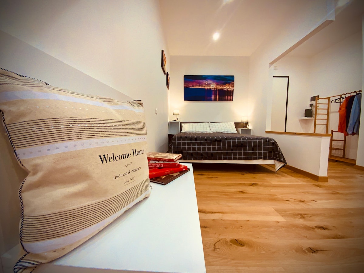 An open space studio is shown, featuring a comfortable bed with a simple, dark plaid cover. A decorative pillow with 'Welcome Home' is placed on a nearby side table. Warm wooden flooring complements the white walls, enhancing the inviting ambiance of the room.