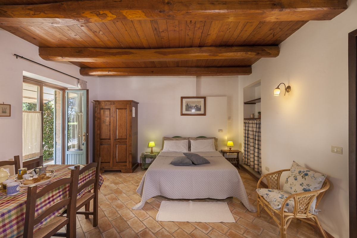 A spacious studio is showcased, featuring a double bed centered beneath a wooden beamed ceiling. An antique wardrobe stands against the wall. A dining table is set with chairs, and natural light enters through large windows, which lead to an outdoor terrace.