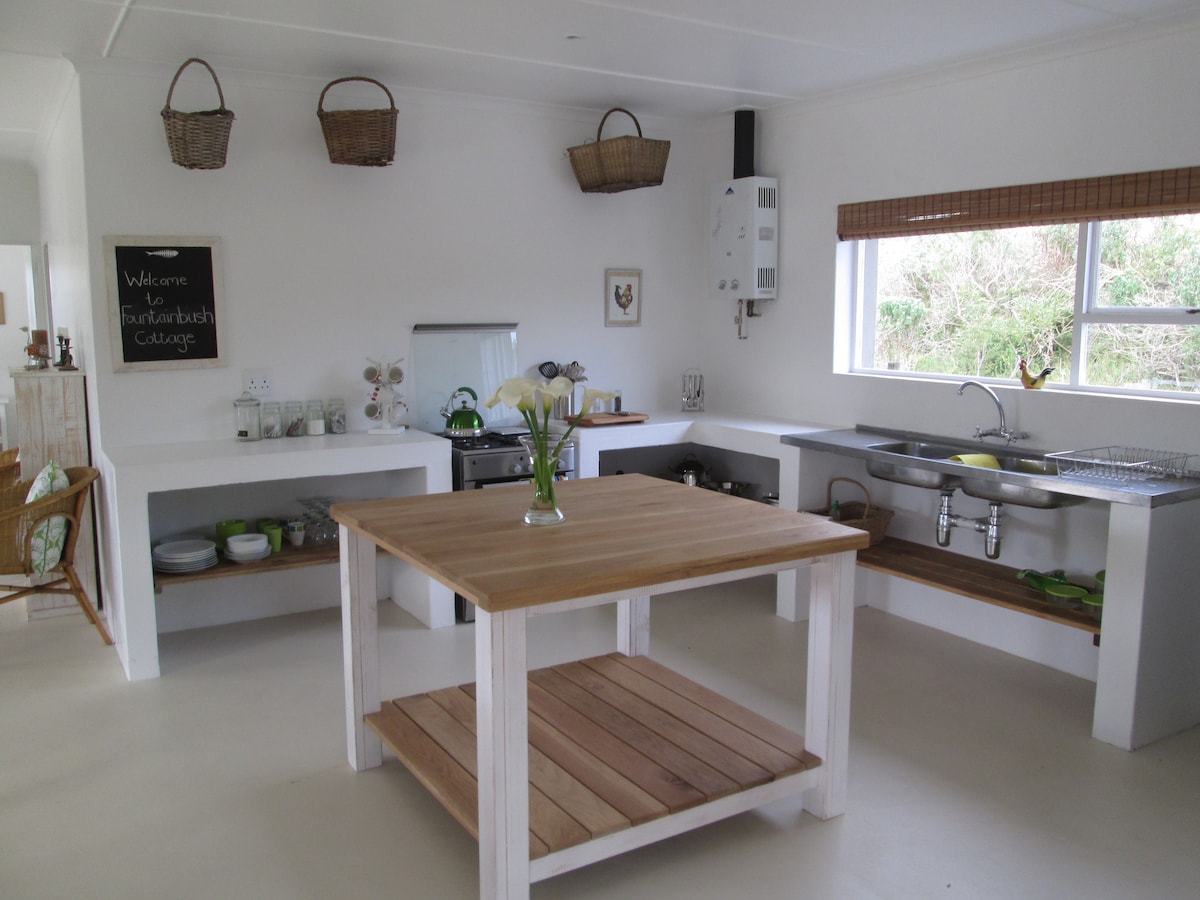 A modern kitchen space features a central wooden island table and practical shelving. Light countertops hold dishware and utensils. Natural light enters through the windows, illuminating the simple design that includes a gas stove and kitchen sink, creating an inviting cooking area.