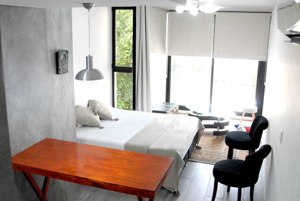 A well-lit studio apartment features a spacious bed adorned with neutral-colored linens. A wooden table sits between a pair of upholstered chairs, while large windows allow natural light to filter through light-colored shades, offering views of greenery outside.