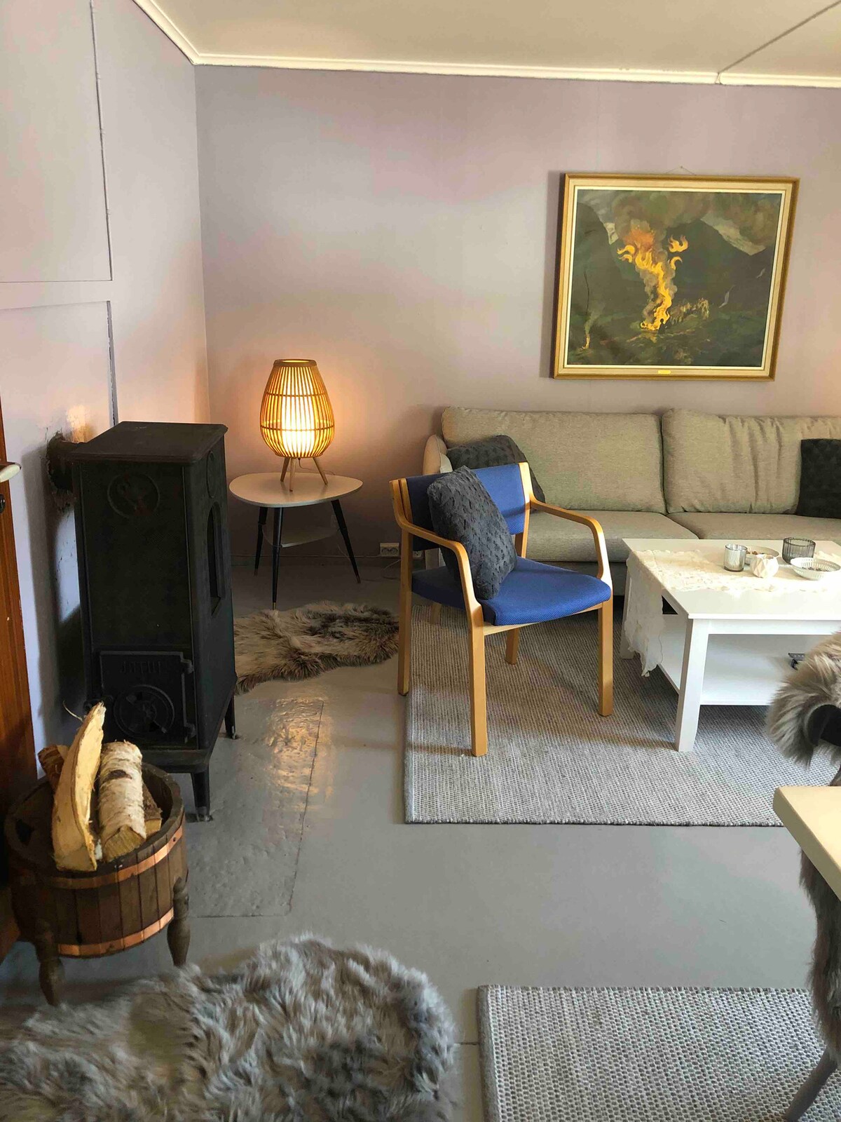 A cozy living room features a soft gray sofa, accompanied by a blue chair. A small table is set with glasses. A warm light is emitted from a lamp beside the sofa. Textured rugs adorn the floor, while a piece of artwork adds color to the wall.