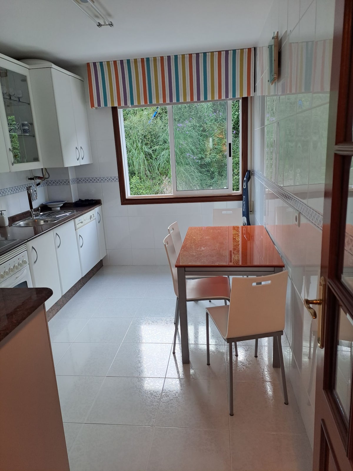 A bright kitchen is equipped with modern cabinetry and a view of greenery through a large window. A wooden table with four chairs is positioned in the space, and additional kitchen amenities are integrated along the walls.