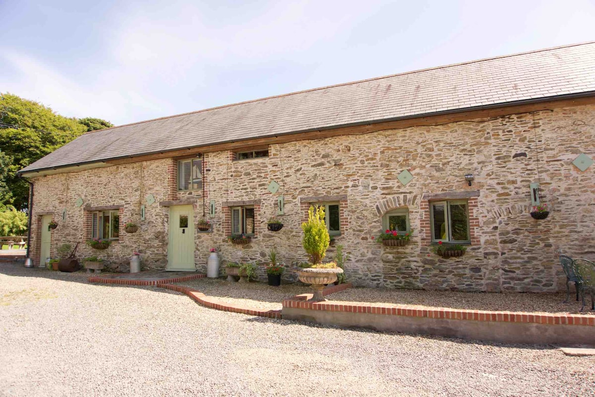 The exterior of a converted stone barn is showcased, featuring a rustic facade with flowering window boxes and decorative stone details. A gravel path leads to the entrance, flanked by potted plants and greenery, offering a warm and inviting approach to the property.