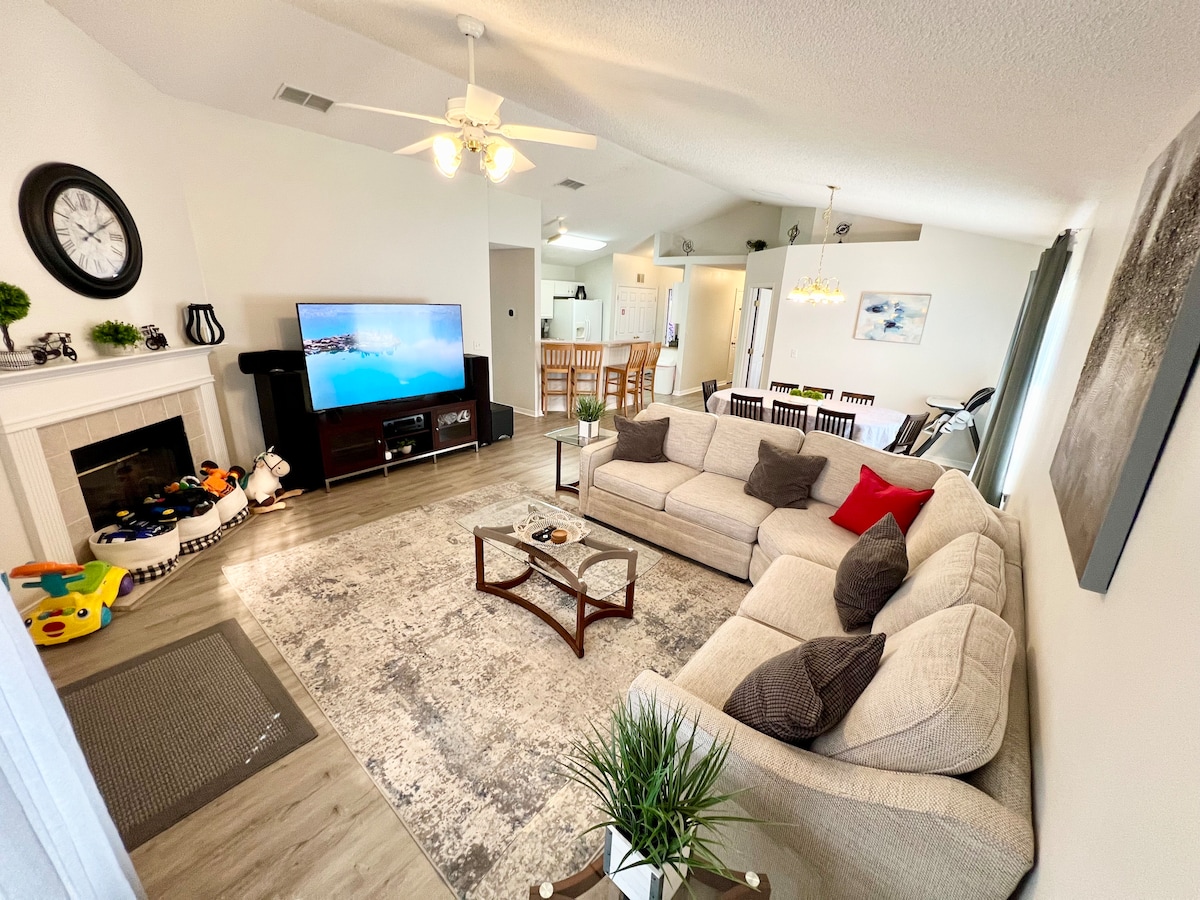 The living room features a spacious sectional sofa with accent pillows, a glass coffee table, and a large TV on a dark wood console. Natural light enters through windows, and a ceiling fan adds to the comfort of the relaxed atmosphere.