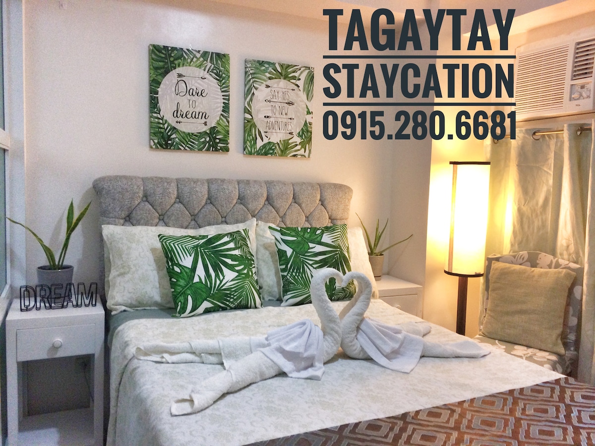 A cozy bedroom is presented with a plush headboard and adorned with green tropical-themed cushions. Light-colored bedding is complemented by decorative towels arranged in the shape of swans. Artwork featuring foliage adds a relaxing touch to the space, enhanced by soft lighting from a standing lamp.