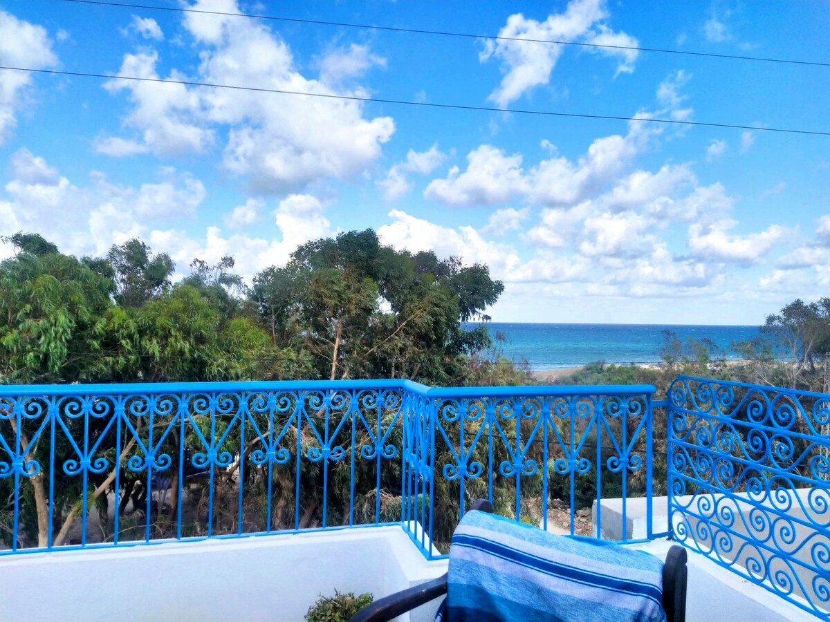 A rooftop terrace showcases a vibrant blue railing overlooking a scenic view of the sea and lush greenery. A single chair with a striped cushion is visible, inviting relaxation while enjoying the expansive sky filled with soft clouds.