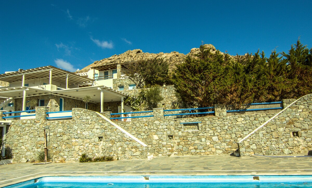 A spacious stone structure is depicted with multiple levels and an expansive terrace overlooking a swimming pool. Lush greenery and natural rock formations rise in the background, complementing the clear blue sky above.