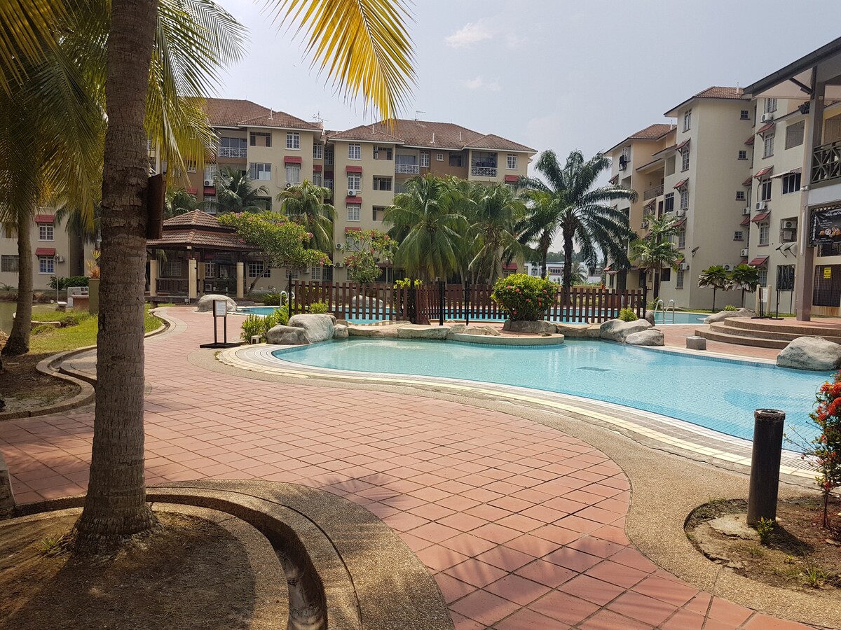 The image showcases a spacious outdoor swimming pool surrounded by lush greenery and palm trees. A curved pathway leads to the pool area, with lounge chairs and shaded spots available for relaxing. Nearby, a gazebo offers additional seating and a communal gathering place.