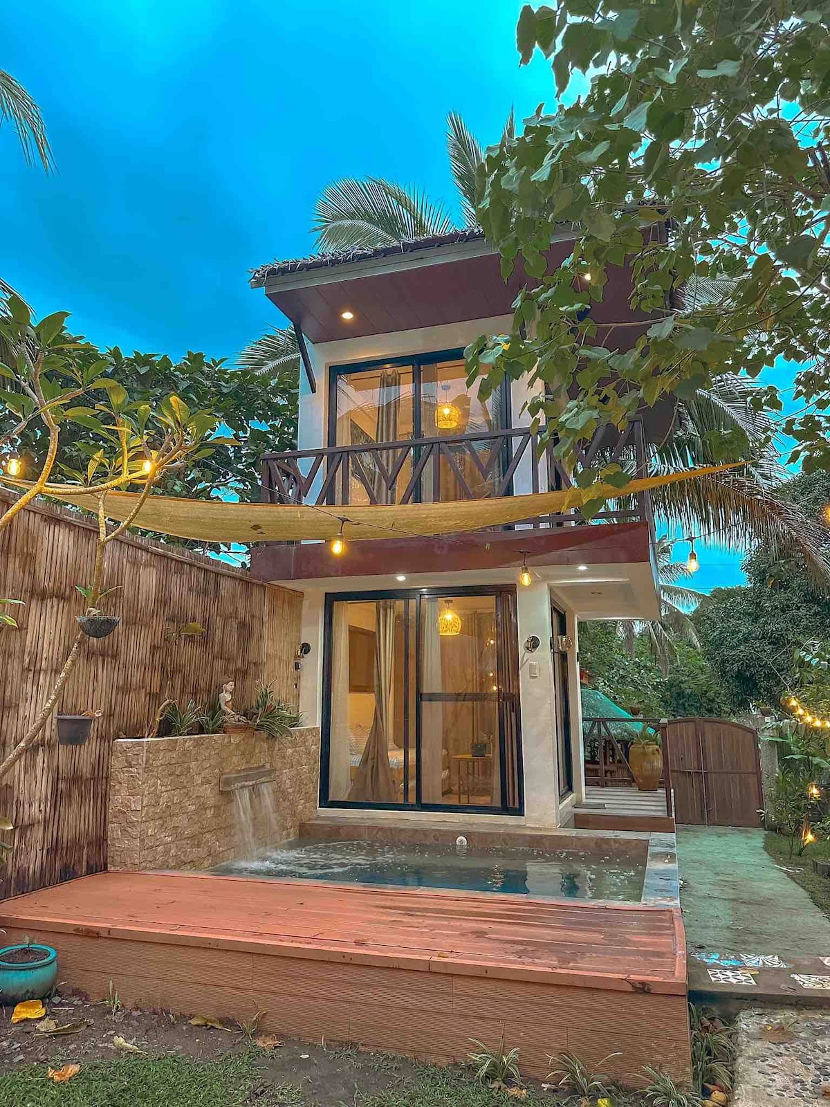 A two-story rustic house is surrounded by lush greenery, featuring a wooden deck that leads to a small dipping pool. Warm outdoor lighting casts a soft glow, while large glass doors provide a view into the interior's inviting space.