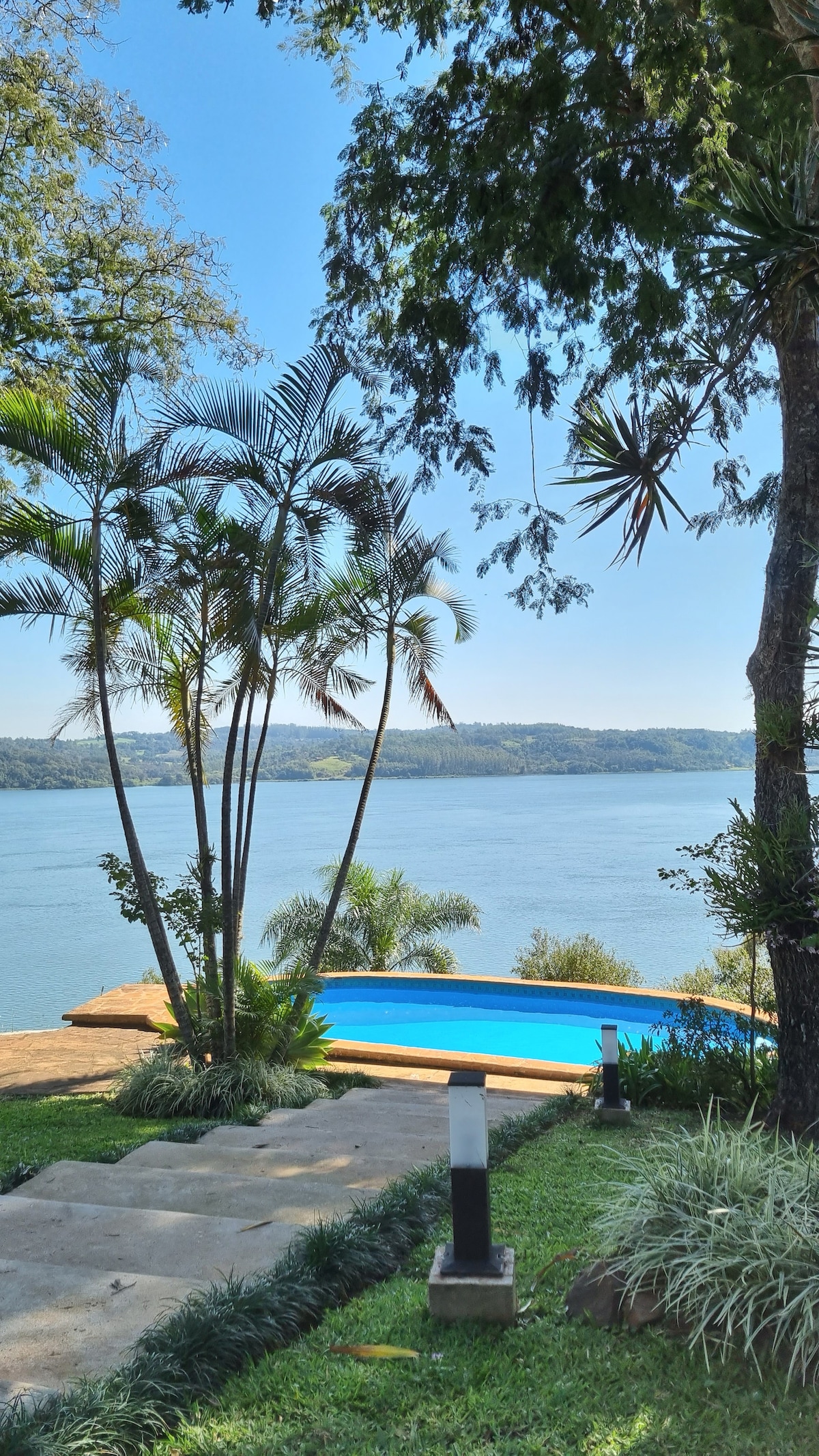 A scenic view of a swimming pool overlooks a tranquil body of water, framed by lush greenery and palm trees. The pool area features a wooden deck, seamlessly blending the natural surroundings with a relaxing outdoor retreat.