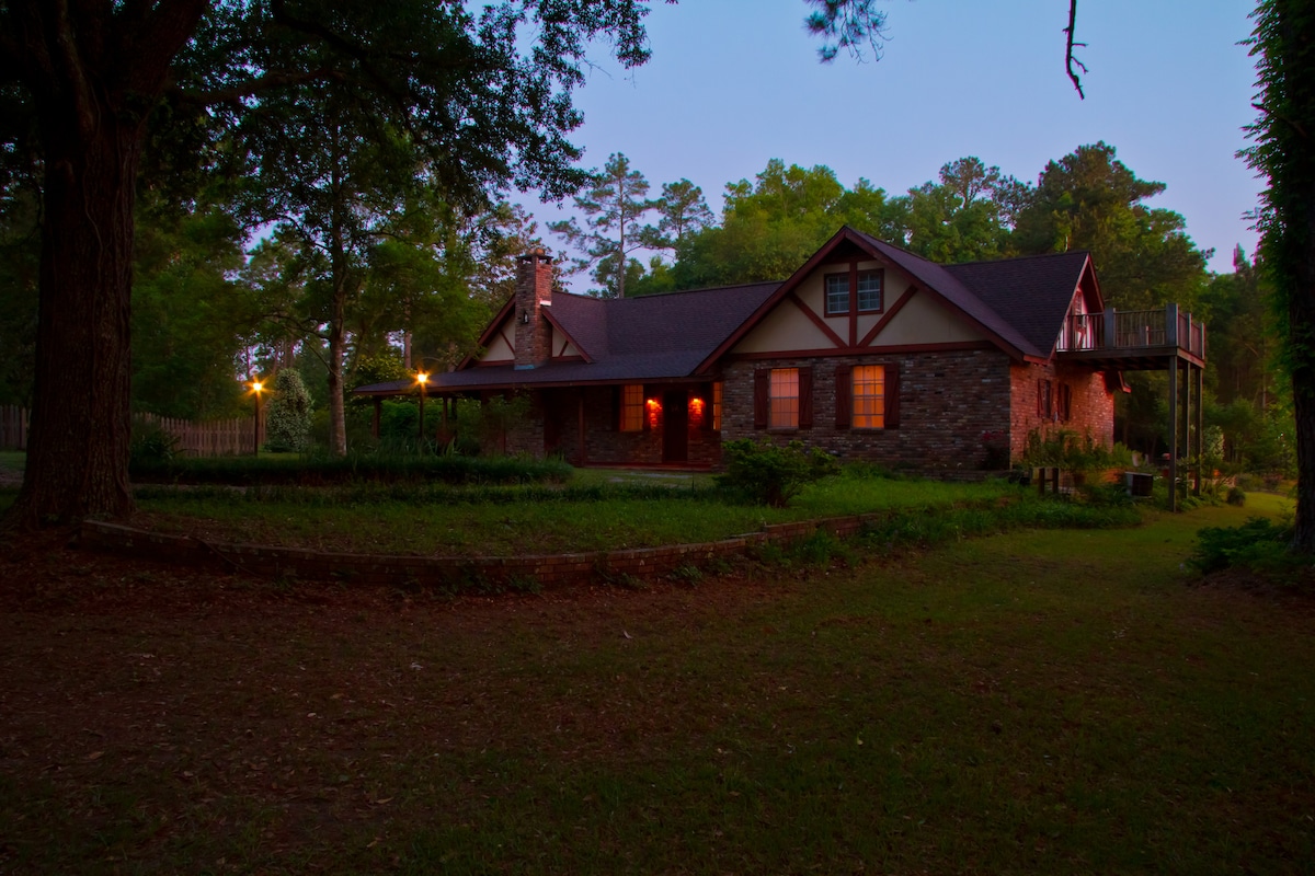 The exterior of a charming two-story country home is set amidst lush greenery, with warm lights emanating from its windows. A stone façade enhances its rustic appeal, while a spacious porch encourages outdoor relaxation. Trees surround the property, creating a serene atmosphere.