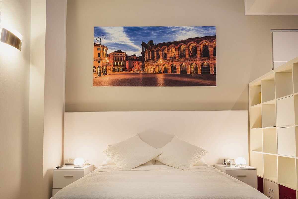 A bright sleeping area features a spacious bed with two white pillows and a light-colored headboard. Above the bed, a large panoramic image of the Arena is displayed. Soft lighting from lamps on either side of the bed enhances the room's ambiance.