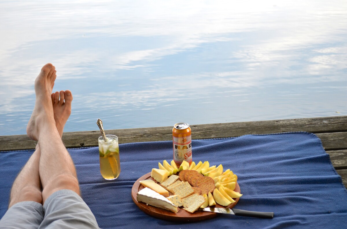 A wooden dock extends over a calm lake, with a blue blanket spread across it. A wooden platter holds a variety of crackers, cheese, and sliced apples. A drink is placed nearby, reflecting the peaceful atmosphere of the lakeside setting.