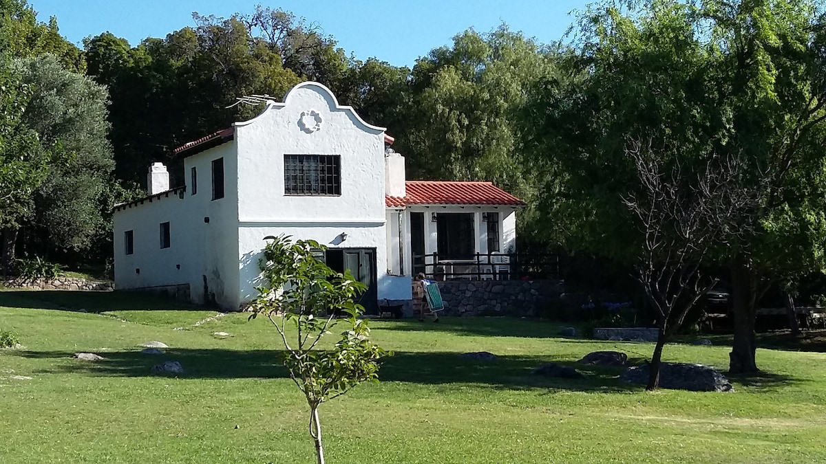 A charming white house features a red-tiled roof and large windows. The surrounding area is composed of a green lawn dotted with rocks and trees, providing a serene outdoor environment. The house is located amidst natural greenery, enhancing the inviting landscape.