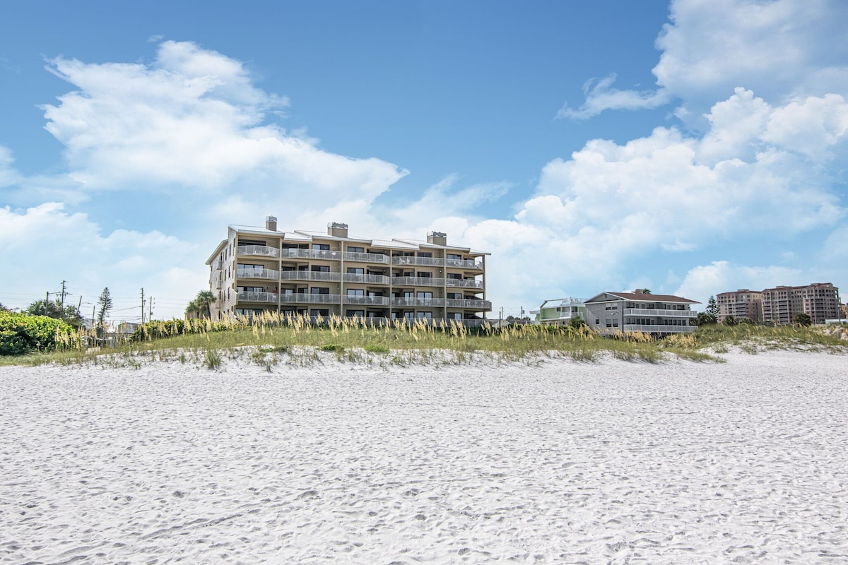 The beachfront condominium is viewed from the soft white sand. The three-story building features multiple balconies, surrounded by coastal grasses and the clear blue sky above. Nearby, additional structures hint at local amenities, providing context to the beachside setting.