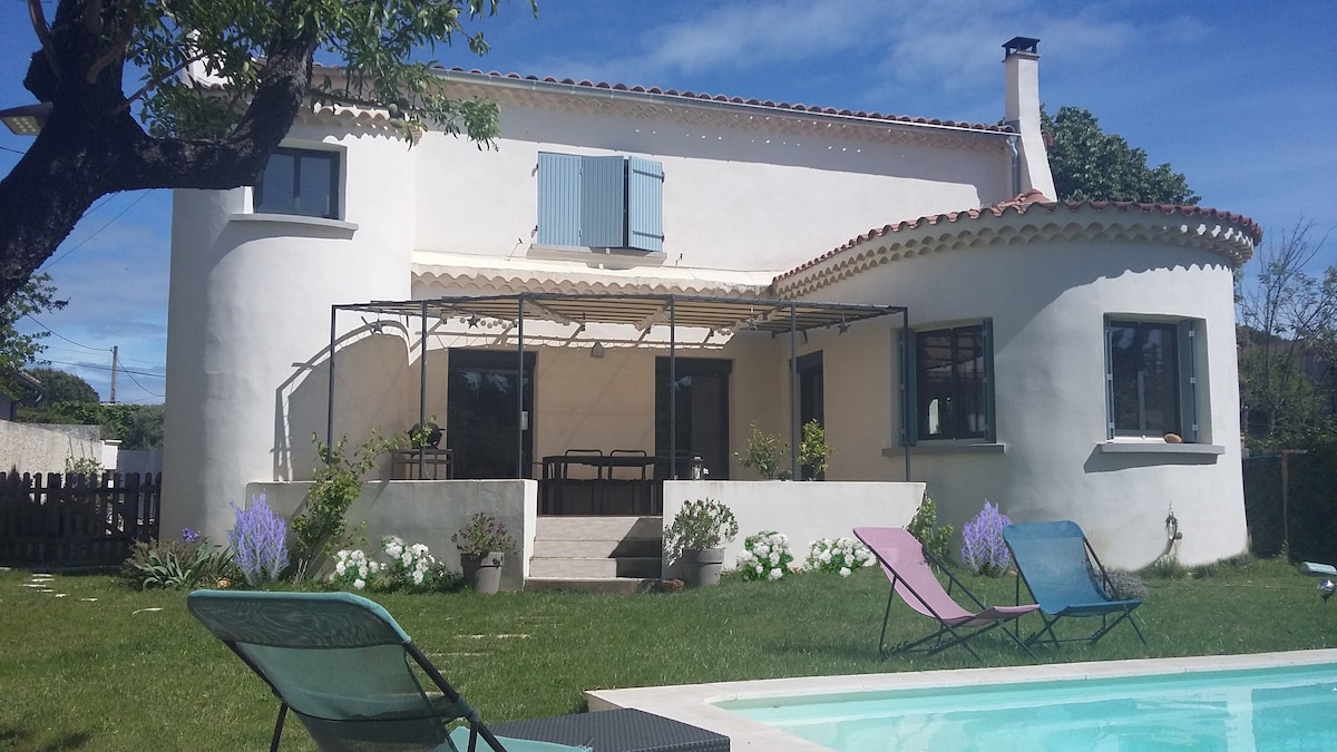 The exterior of a charming home is highlighted, featuring round architectural elements and large windows. A shaded terrace with a dining area is visible, surrounded by a well-kept garden with white flowers and green grass. A swimming pool and loungers provide outdoor relaxation options.
