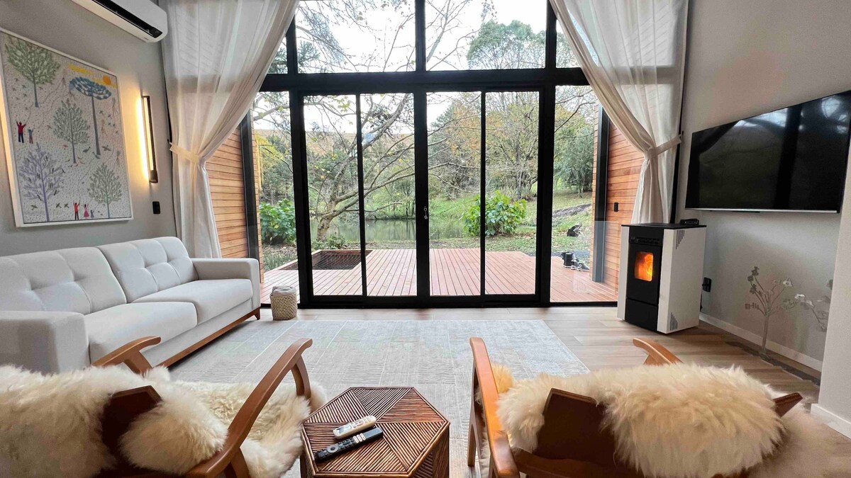 A cozy living area features a comfortable white sofa and two wooden armchairs adorned with soft throws. Large glass doors open to a scenic view of the garden, allowing natural light to fill the space. A contemporary stove adds warmth and a touch of modern design.