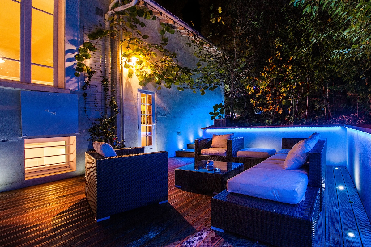 An outdoor terrace is illuminated with soft blue lighting, showcasing comfortable seating arrangements with cushioned sofas and a coffee table. Lush greenery surrounds the space, enhancing the tranquil environment. Warm light filters from nearby windows, creating a welcoming atmosphere for relaxation.