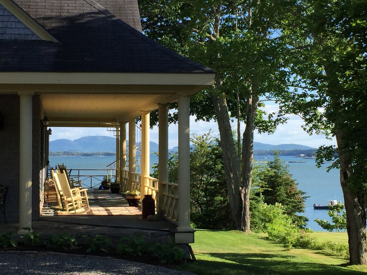 A charming porch area is visible, featuring rocking chairs and expansive views of the ocean. Lush greenery surrounds the space, with distant mountains and boats gently floating on the water. Soft natural light enhances the serene atmosphere, inviting relaxation amidst nature.