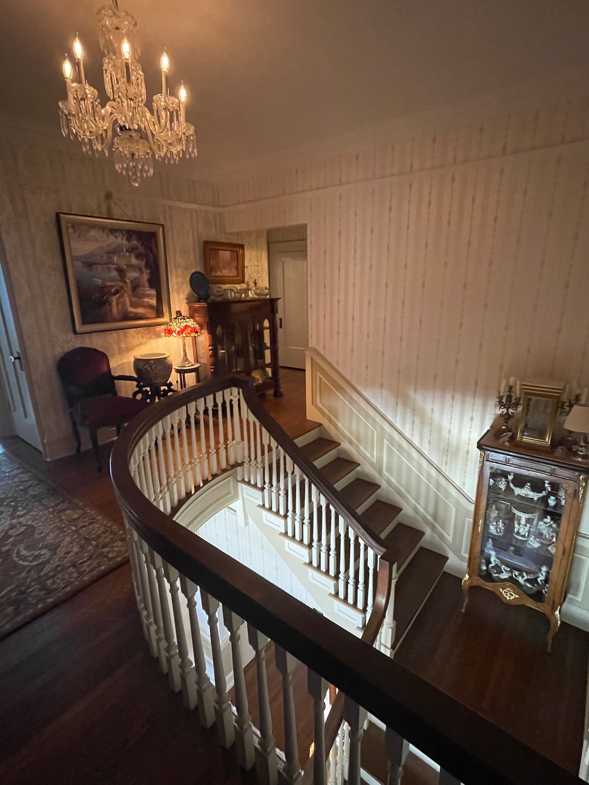 An elegant staircase with a curved design leads to the upper level. A chandelier hangs above the landing, while a decorative console and armchair are positioned nearby. The walls are adorned with subtle wallpaper, and light streams in from a doorway unseen.