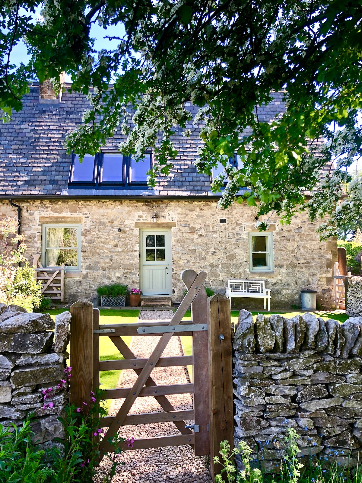 A charming stone cottage is framed by the lush greenery of surrounding trees. A welcoming wooden gate leads to a gravel pathway that guides to the cottage's entrance, showcasing a mix of rustic stonework and modern window designs. A small garden area is visible, adding to the inviting atmosphere.