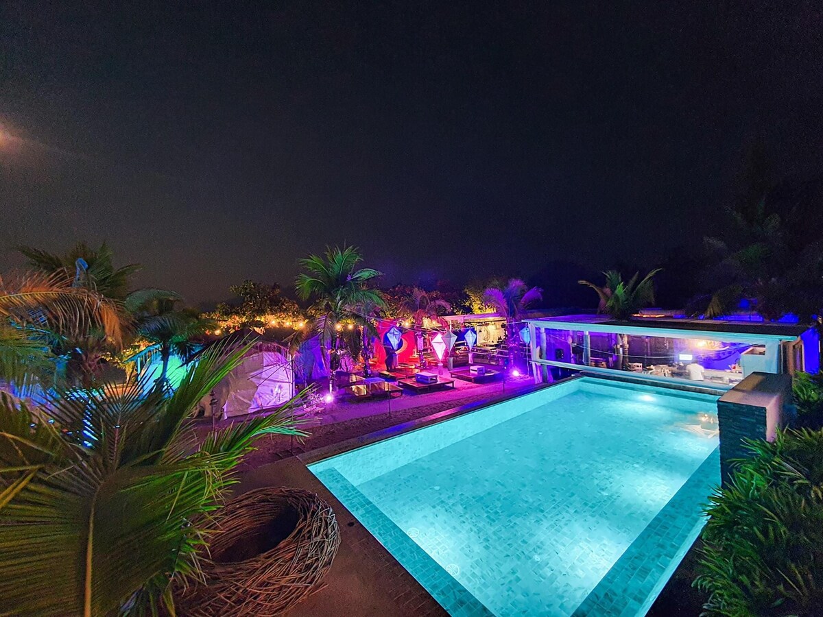 A modern outdoor pool area is illuminated by colorful lighting, creating an inviting atmosphere at night. Surrounding palm trees add lush greenery, while various seating areas and lounge chairs are positioned nearby. The pool's clear water reflects the vibrant lights, enhancing the tranquil setting.