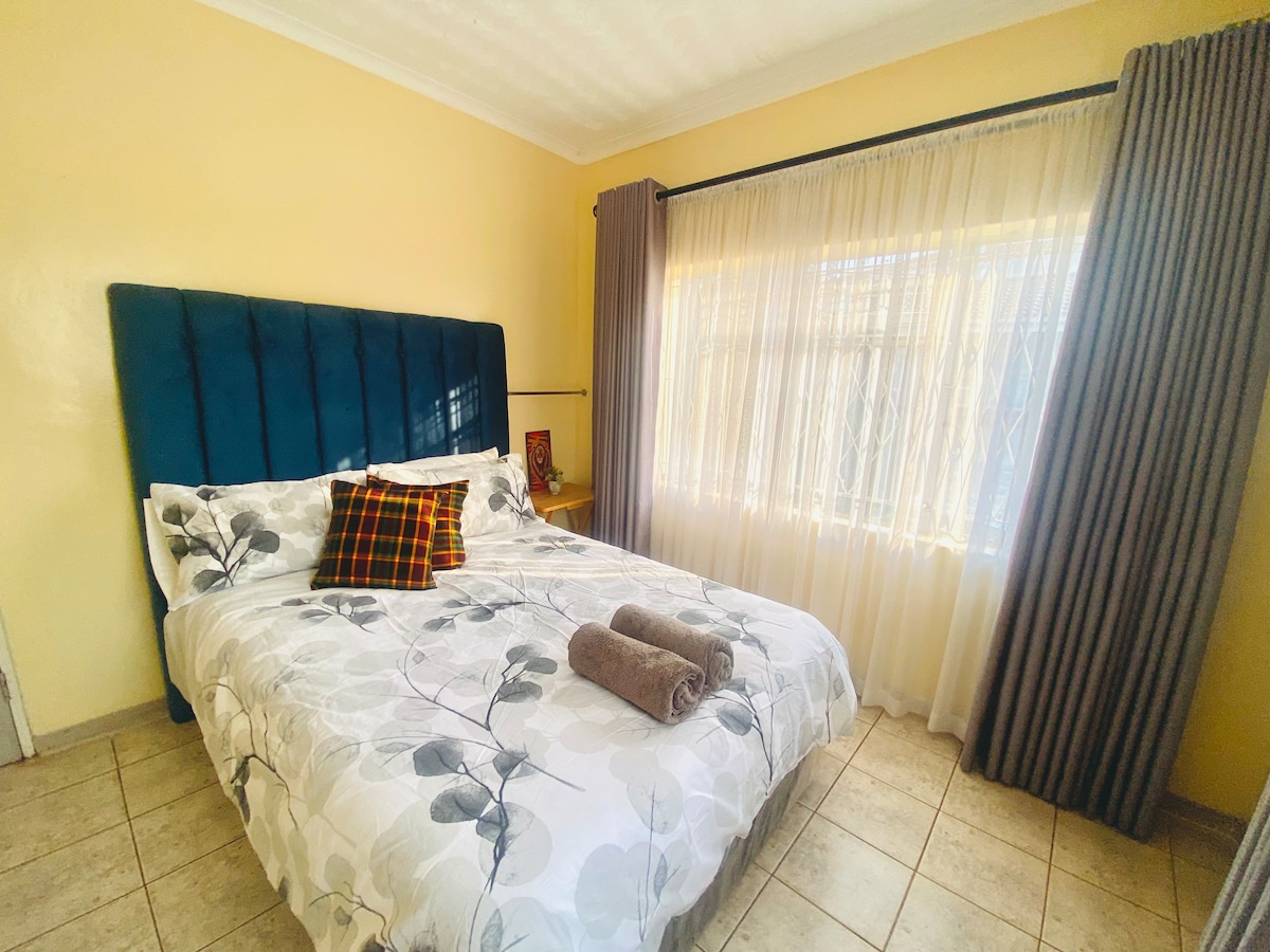 A spacious sleeping area is highlighted by a soft blue headboard and floral bedding. Two neatly rolled towels are placed on the bed. Natural light filters through sheer curtains, complementing the warm yellow wall and tiled floor.