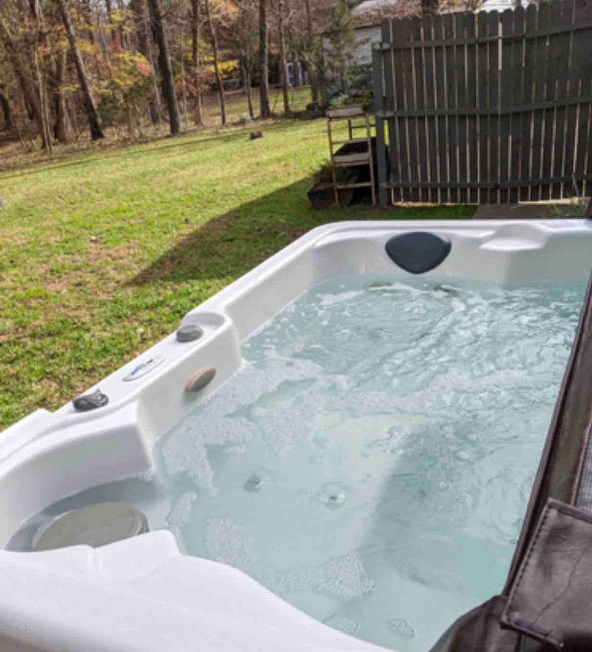 A spacious hot tub filled with clear water is shown, situated on a patio. The surrounding yard features lush green grass, bordered by a wooden fence, enhancing privacy. Trees in the background provide a natural setting.