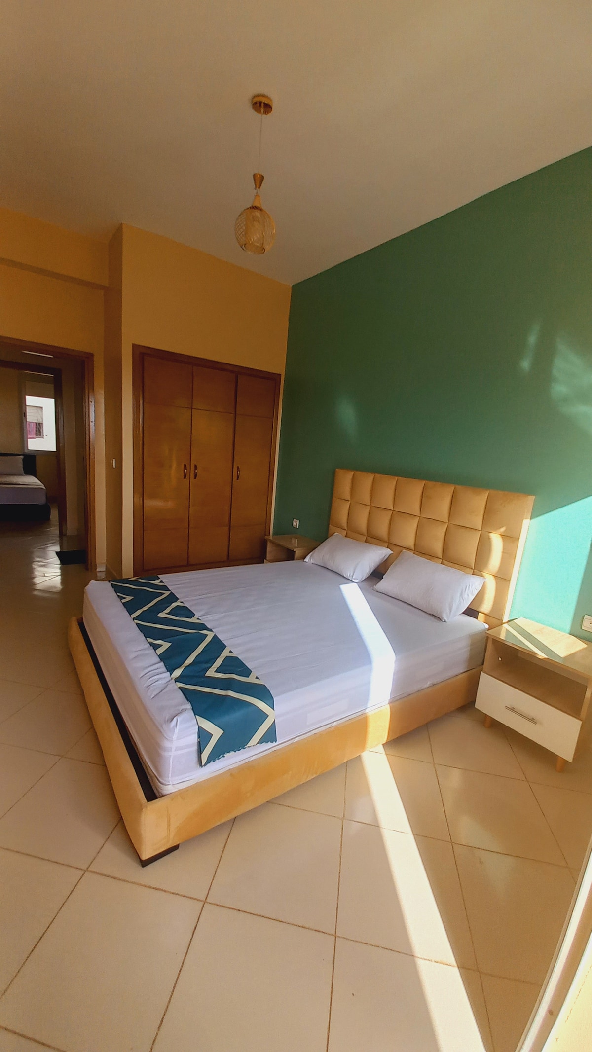 A brightly lit bedroom is presented, featuring a spacious bed with a beige upholstered headboard and crisp white linens. A green accent wall adds a pop of color. Two wooden cabinets are positioned along one wall, enhancing the room's functionality.