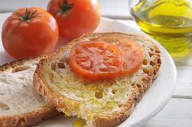 A slice of toasted bread is topped with a fresh slice of tomato, resting on a white plate. Two ripe tomatoes sit nearby, complemented by a small bowl of olive oil. The setting reflects a simple and inviting dining experience.