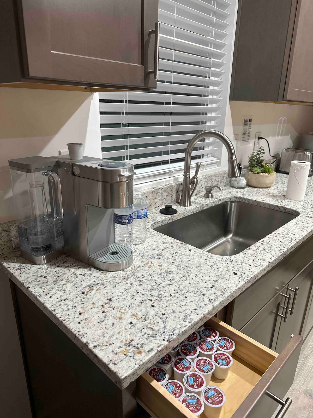 A modern kitchen counter features a sleek coffee maker alongside a sink with a polished faucet. A drawer showcases various coffee pods, while bottles of water are visible on the counter. Natural light filters through a window adorned with blinds.