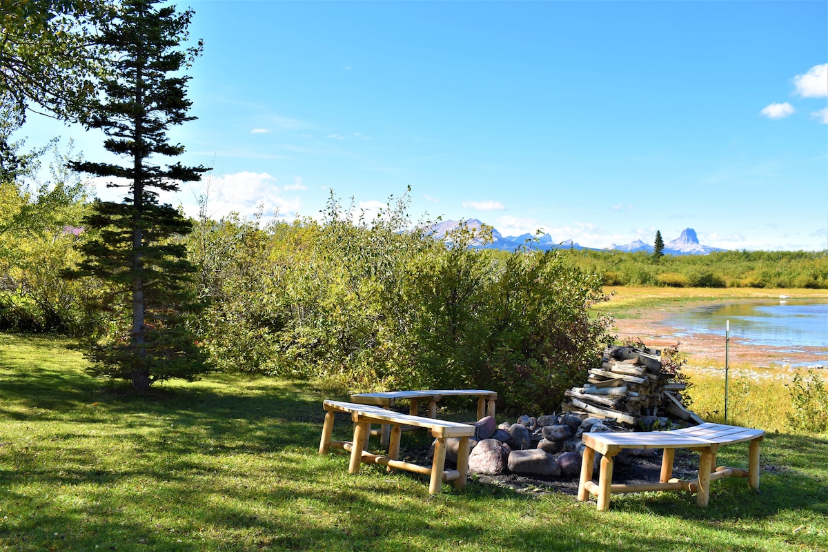 An outdoor gathering area features two wooden benches positioned near a stone fire pit, surrounded by lush greenery and tall grasses. The serene landscape includes a scenic view of a lake and distant mountains under a clear blue sky.