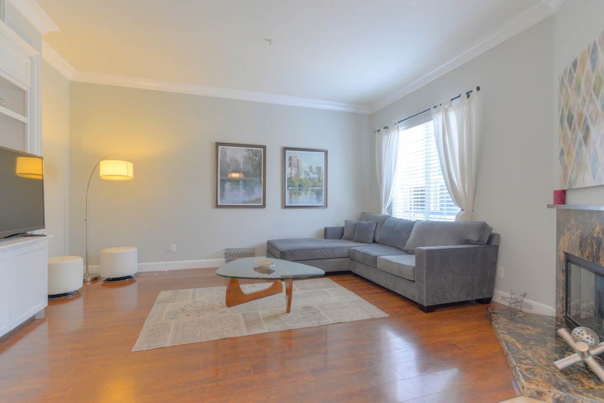 A bright living room features large windows allowing natural light to fill the space. A comfortable gray sectional sofa is positioned near a coffee table, complemented by a decorative area rug. Artwork is displayed on the walls, and a sleek TV unit is visible.