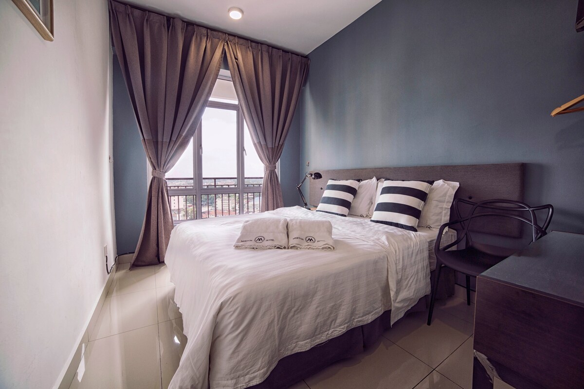 A well-lit bedroom features a spacious bed with soft linens and two decorative pillows. A large window draped with curtains offers views of the city. A bedside table and a minimalist chair complement the clean design of the room.
