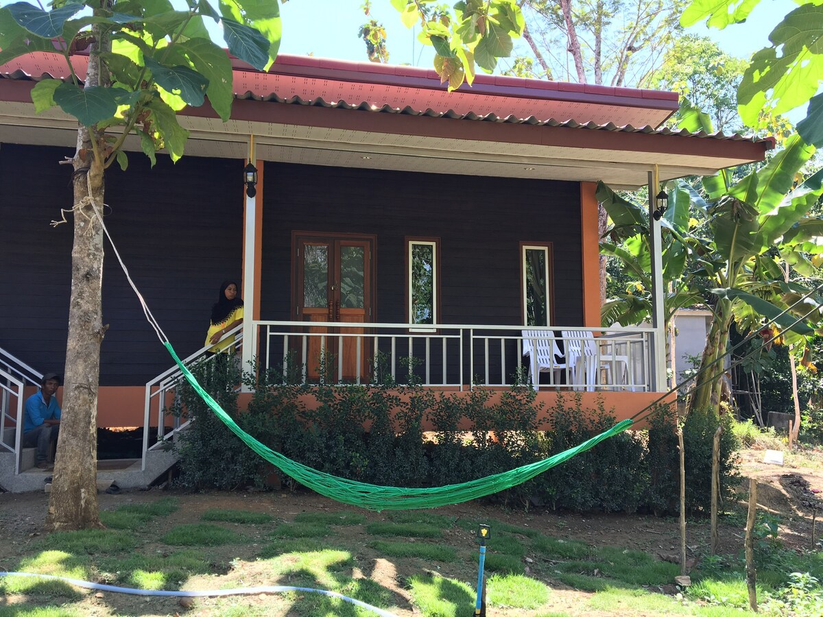 A welcoming exterior of a house is displayed, featuring a red roof and dark walls. A hammock is suspended between two trees, inviting relaxation. The front porch has a few white chairs and multiple windows that allow natural light to enter the space.