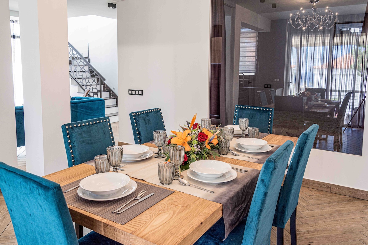 A modern dining area is showcased with a large wooden table surrounded by plush, blue chairs. The table is elegantly set with plates, glassware, and a floral centerpiece. Natural light filters through large windows, enhancing the inviting space.