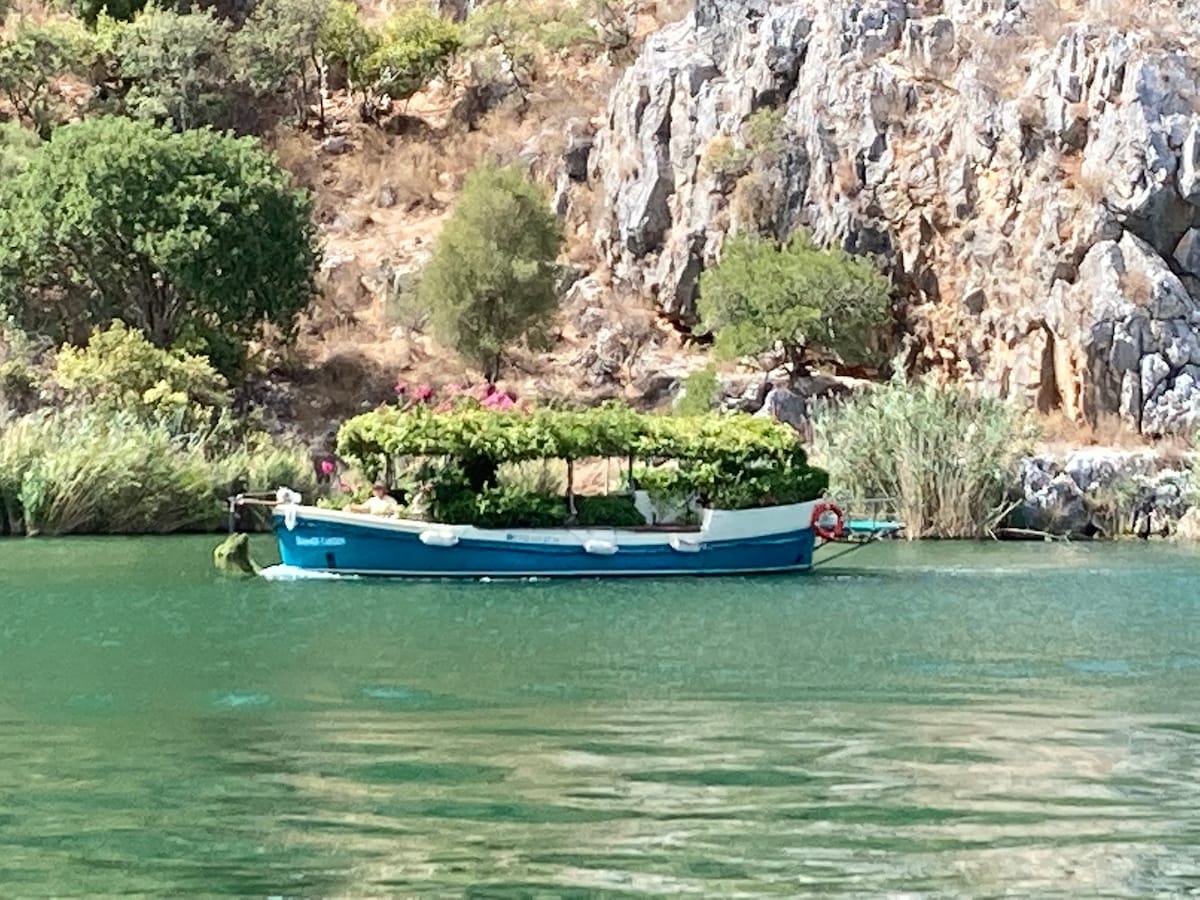 A small blue boat floats quietly on the calm water, surrounded by lush greenery and flowering plants. Among the rocky landscape, trees and shrubs create a natural backdrop, reflecting in the shimmering surface, enhancing the tranquil setting.
