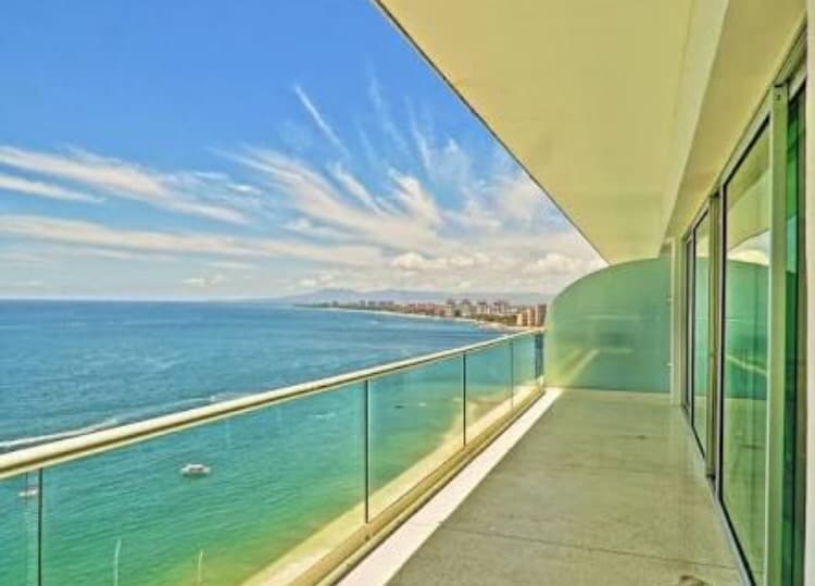A spacious terrace offers sweeping views of the ocean and coastline. Clear glass railings enhance the panoramic perspective while allowing natural light to fill the space. The blue water and sky create a serene backdrop, complemented by distant buildings along the shore.