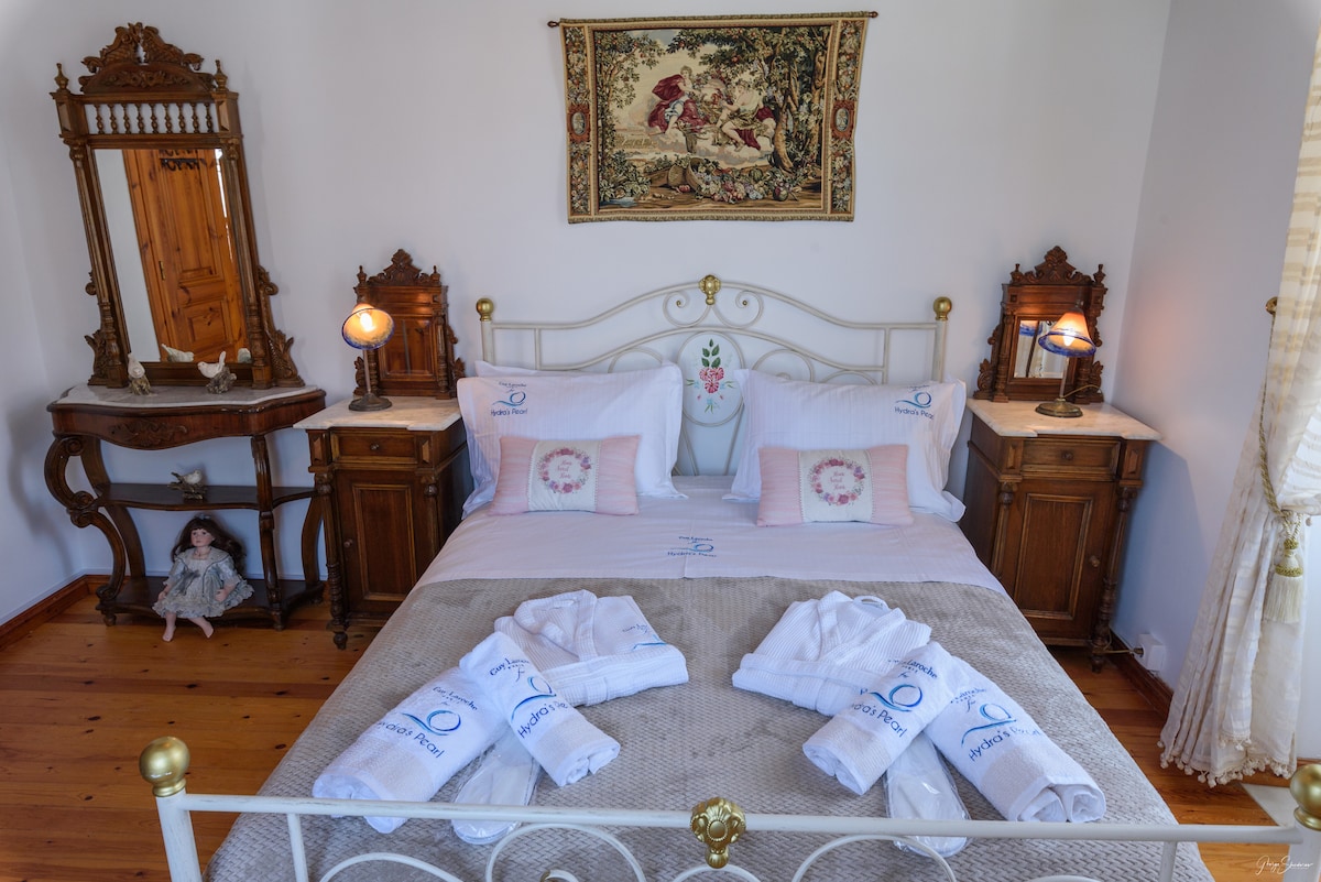 A well-furnished sleeping area features a spacious bed adorned with soft linens and decorative pillows. Two bedside lamps illuminate the space, complemented by antique wooden furniture. Plush towels are neatly arranged at the foot of the bed, enhancing the room's inviting ambiance.