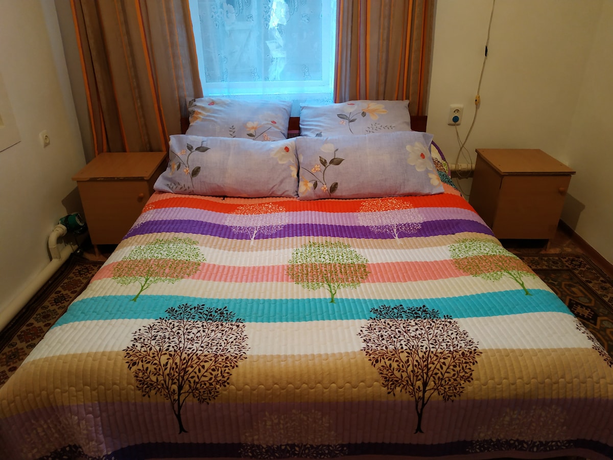 A spacious bed covered with a colorful quilt featuring tree designs is positioned against a window dressed with light curtains. Two nightstands are placed on either side of the bed, providing convenient space for personal items.