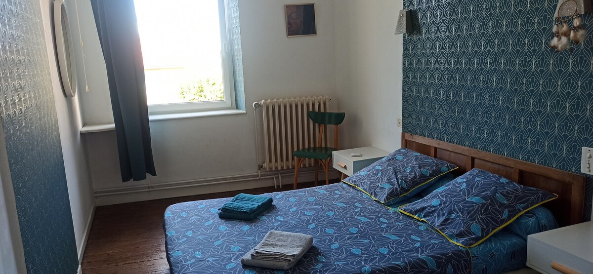 A bedroom is presented with a spacious bed covered in blue patterned linens. Natural light enters through a window, providing a soft glow. A green chair and a small side table are positioned by the window. A radiator is visible against the wall, along with decorative wall art.