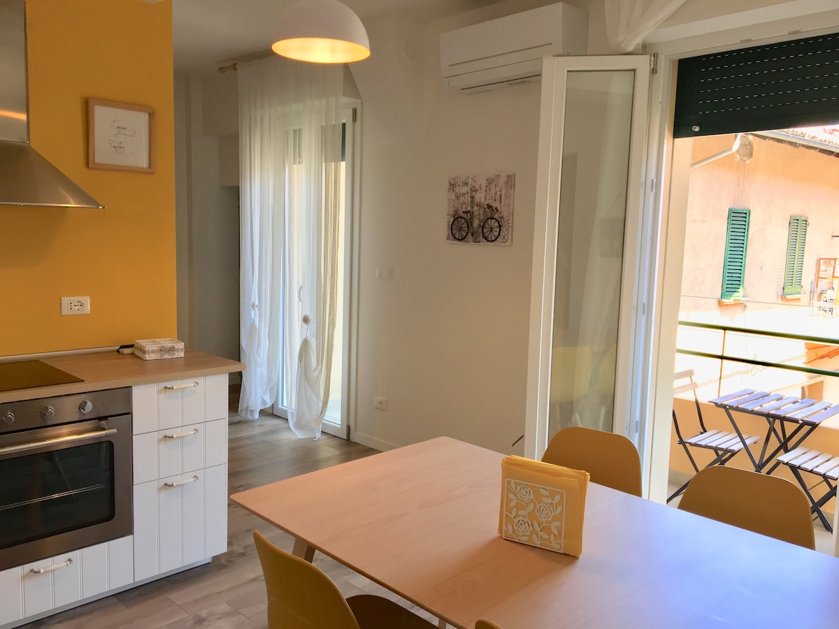 A modern kitchen area is displayed, featuring a bright yellow accent wall. A spacious dining table is surrounded by several chairs, while sliding glass doors open to a small balcony, allowing natural light to fill the room. The kitchen includes an oven and stylish cabinetry.