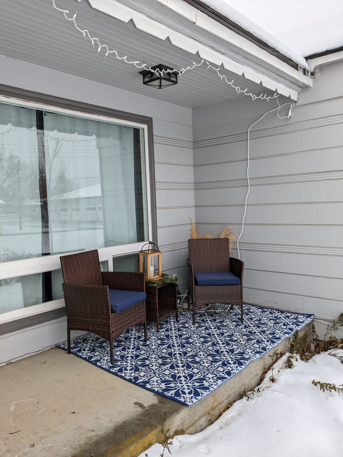 A covered porch features two wicker chairs with blue cushions positioned around a small side table. A lantern is placed atop the table, complementing the decorative blue and white rug. Snow covers the ground, creating a serene winter scene.