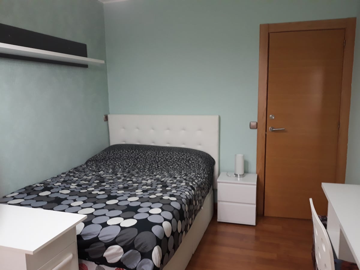 A well-lit room features a spacious bed dressed in patterned bedding, complemented by two nightstands and a desk with a chair. A light green wall adds a fresh touch, while a wooden door provides access to the room. A shelf above the bed offers additional storage.