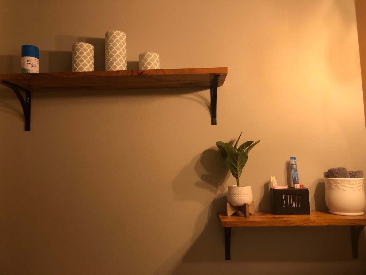 Two wooden shelves are mounted on a wall, displaying decorative items. On the upper shelf, three patterned candles and a blue container are visible. The lower shelf features a small plant, a box labeled 'STUFF,' and toiletries, with a towel set placed nearby.