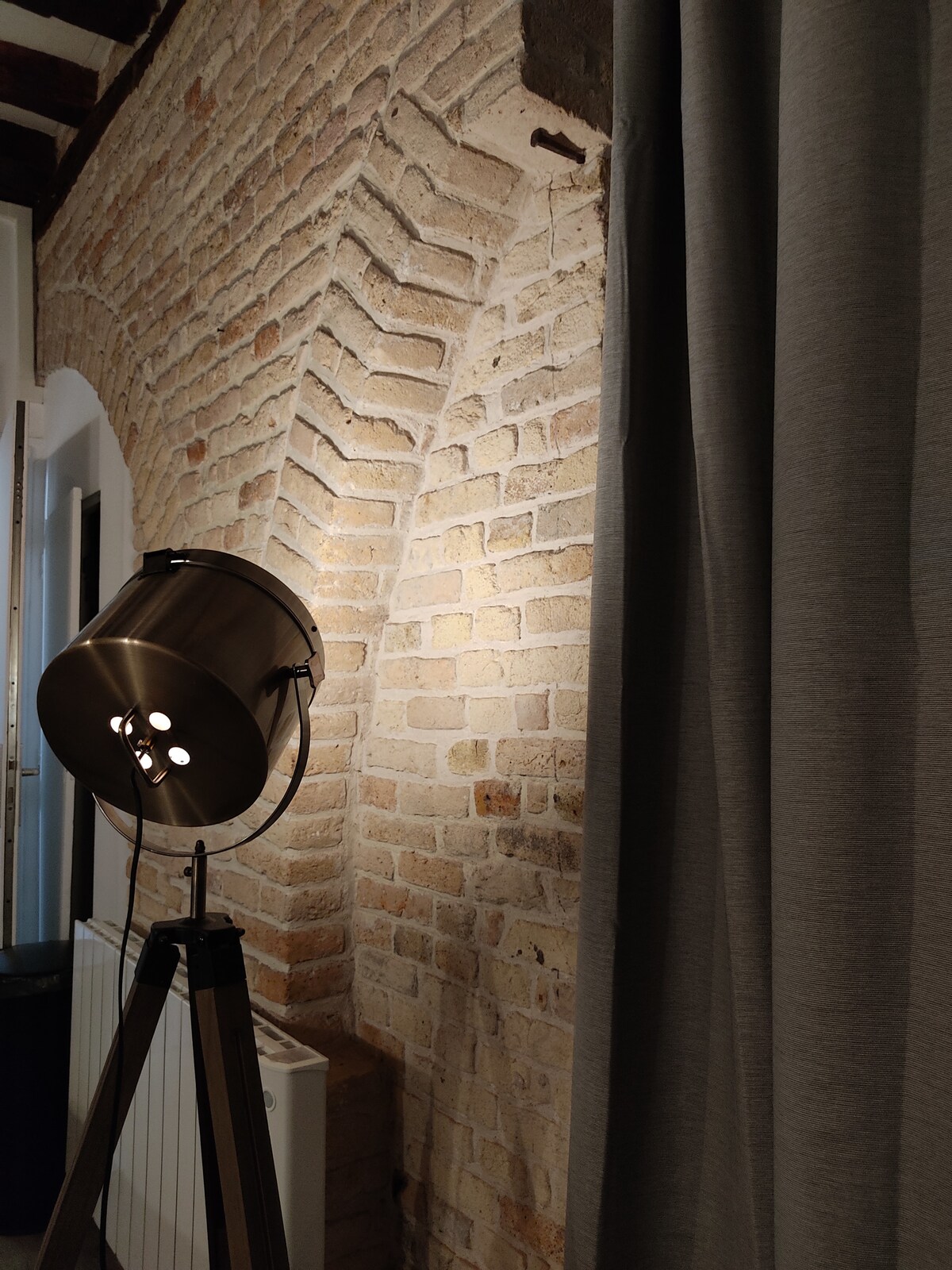 An exposed brick wall is illuminated by a sleek floor lamp, highlighting the textured surface. Grey curtains drape beside the wall, and a radiator is visible in the background, adding to the studio's modern aesthetic. The interplay of light and materials creates a cozy ambiance.