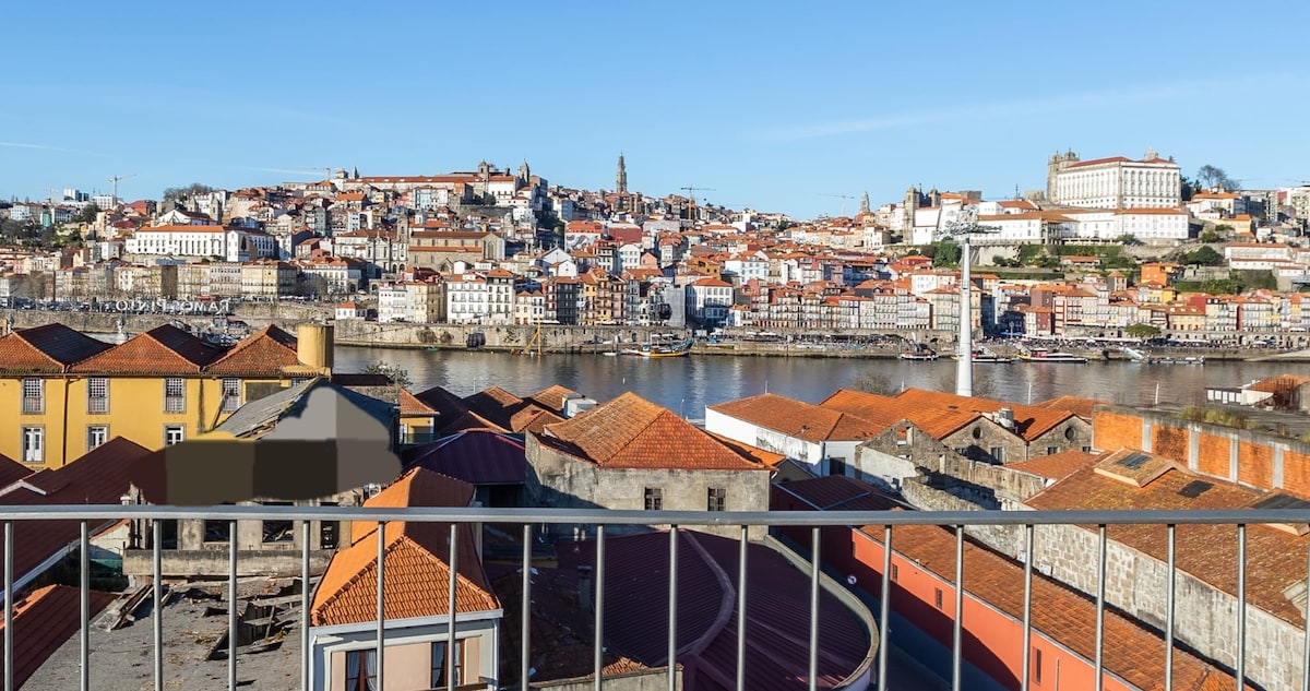 A panoramic view of the historic city of Porto is captured from the terrace, showcasing a layout of colorful houses and terracotta rooftops along the riverbank. The skyline features various architectural styles, with notable landmarks positioned against a clear blue sky.