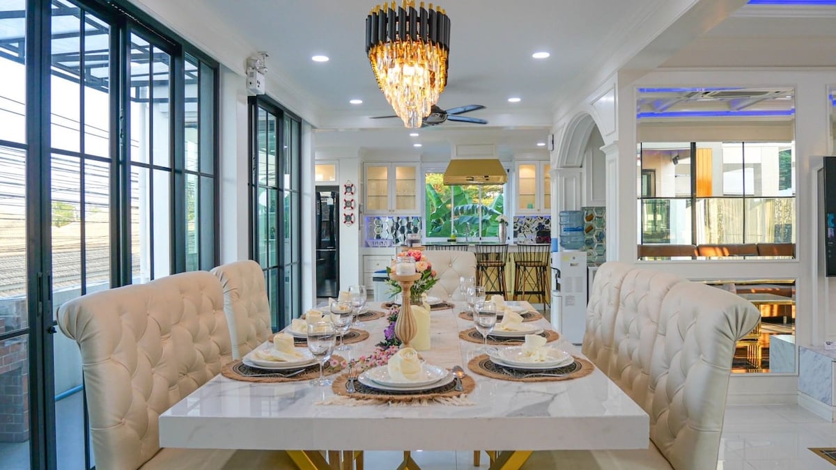 A bright dining area features a large marble table set for six, surrounded by upholstered chairs. Natural light enters through expansive glass windows, enhancing the elegant chandelier above. The room exudes a modern design with neutral tones and carefully arranged table settings.